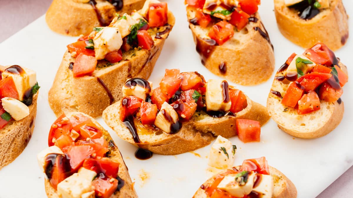 Several slices of gluten-free bruschetta with tomato, mozzarella and a balsamic glaze drizzle on a marble platter.