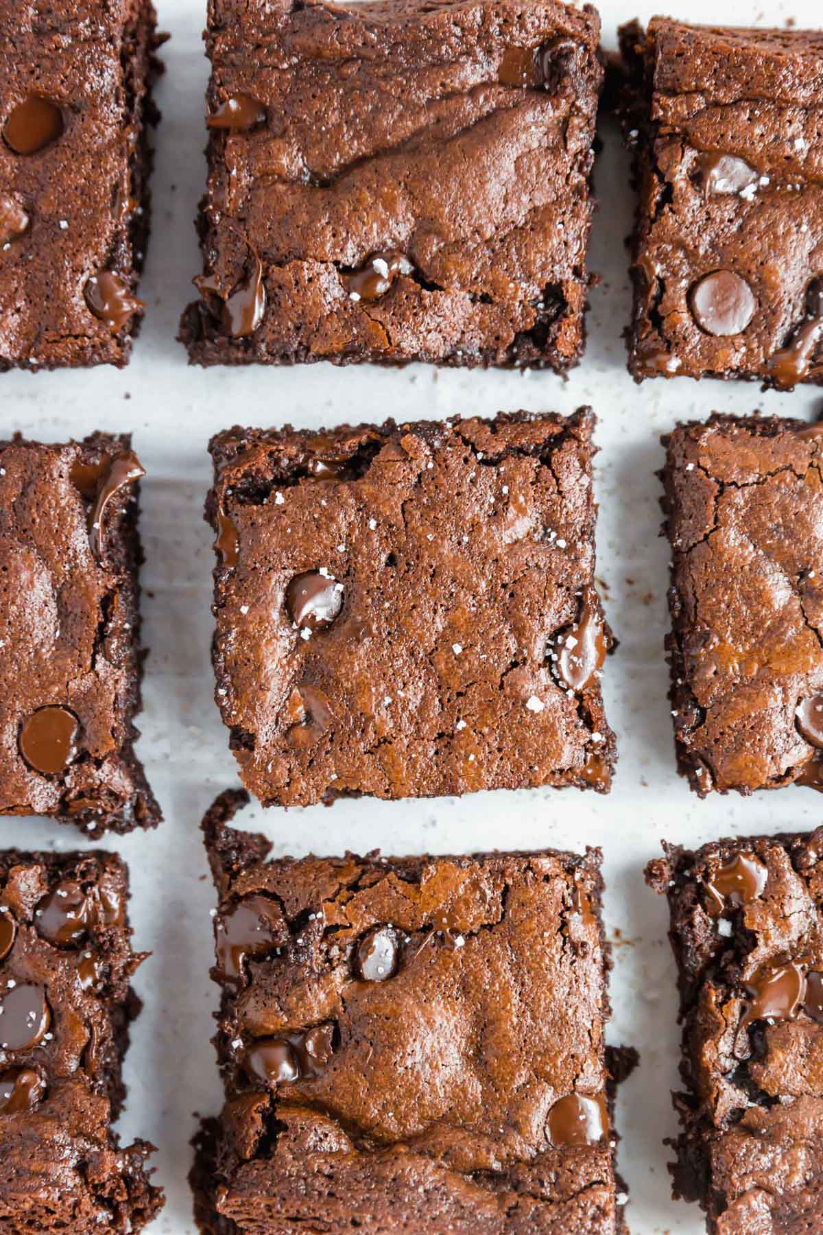 Nine chocolate brownies with chocolate chips are arranged in a grid on a white surface. The brownies have a rich, fudgy texture and a slightly cracked top, with some melted chocolate chips visible throughout.