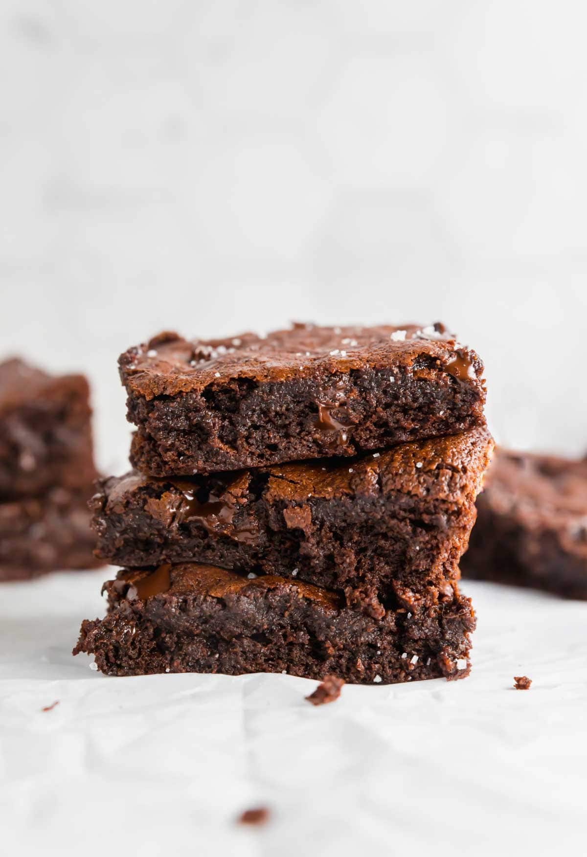 A stack of three gooey chocolate brownies with a crispy top, placed on a white surface. More brownies are visible blurred in the background.