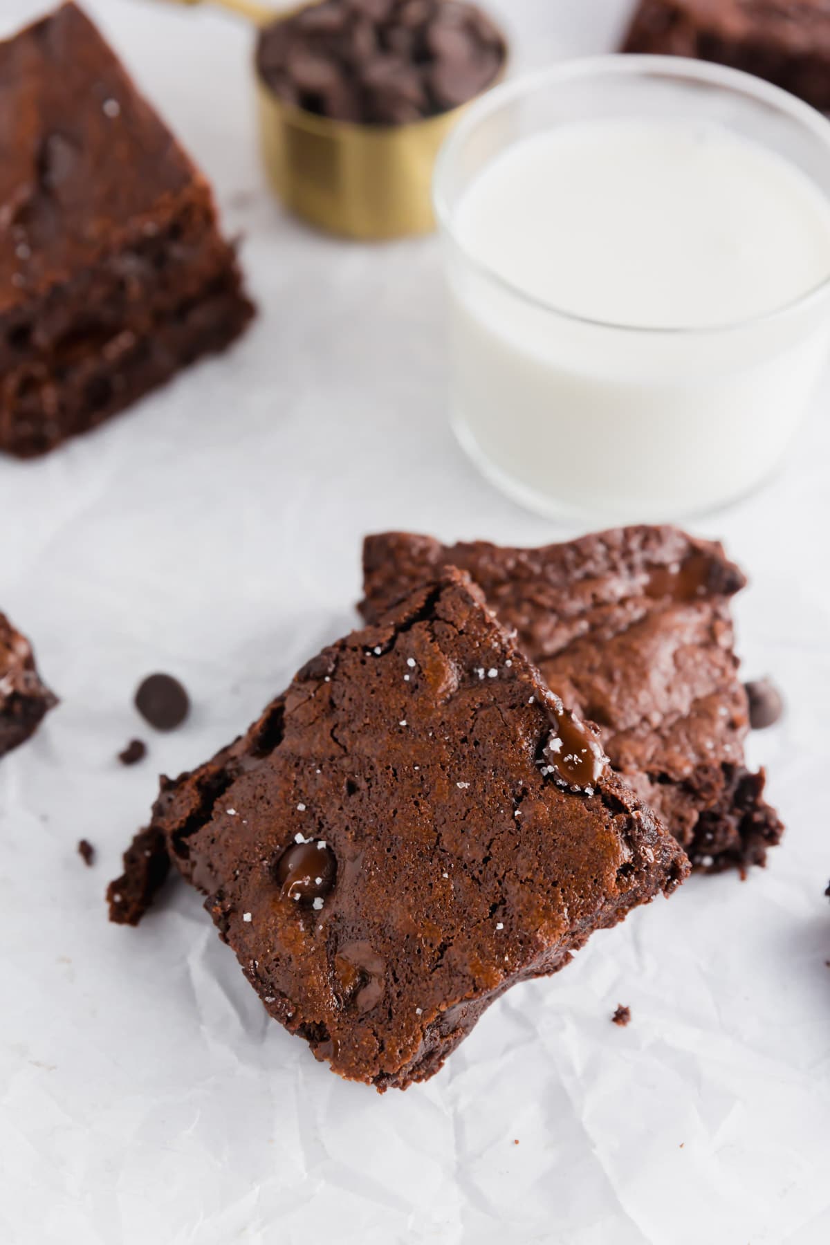 Two fudgy chocolate brownies with chocolate chips are stacked on white parchment paper. A glass of milk and a measuring cup with chocolate chips are in the background.