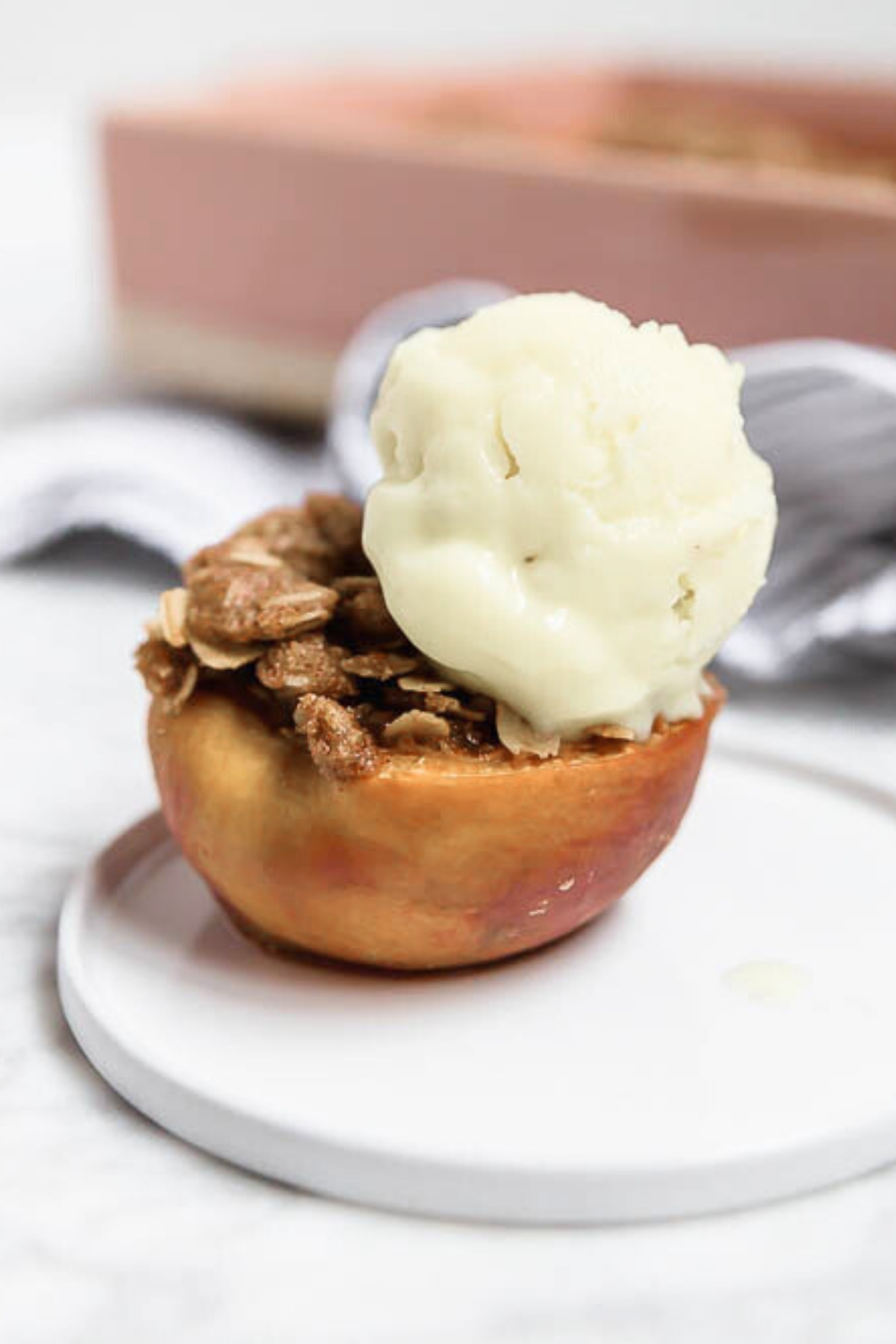 A Baked Peach half with Gluten-Free Streusel is filled with oat crumble and topped with a scoop of vanilla ice cream, served on a white plate against a softly blurred background.