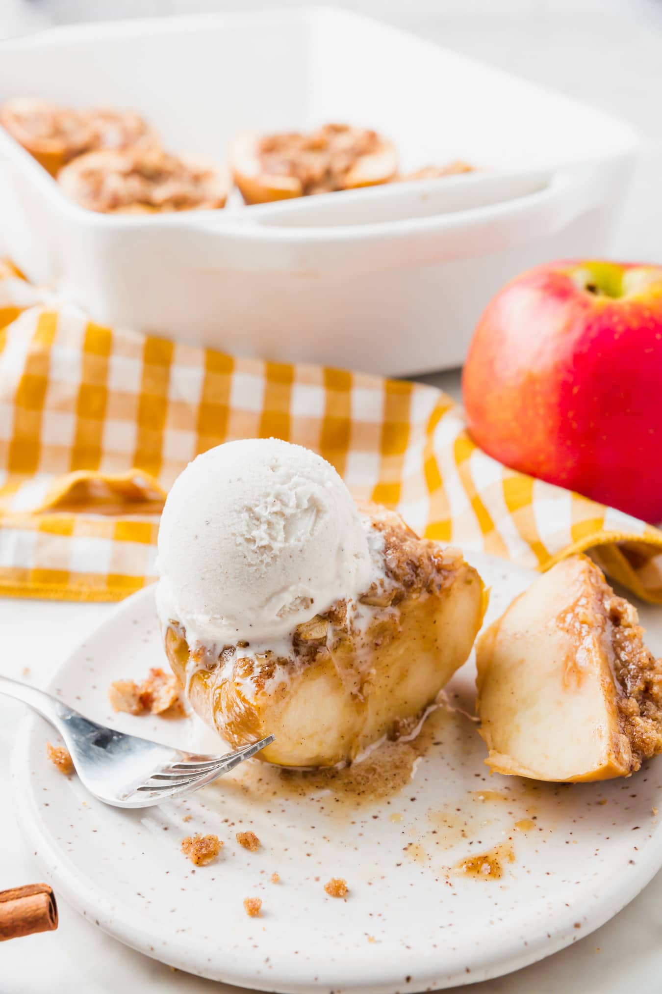 A baked stuffed apple half topped with vanilla ice cream and crumbly streusel sits on a white plate, with a fork and crumbs nearby. A whole apple, yellow-checked cloth, and baking dish are in the background.