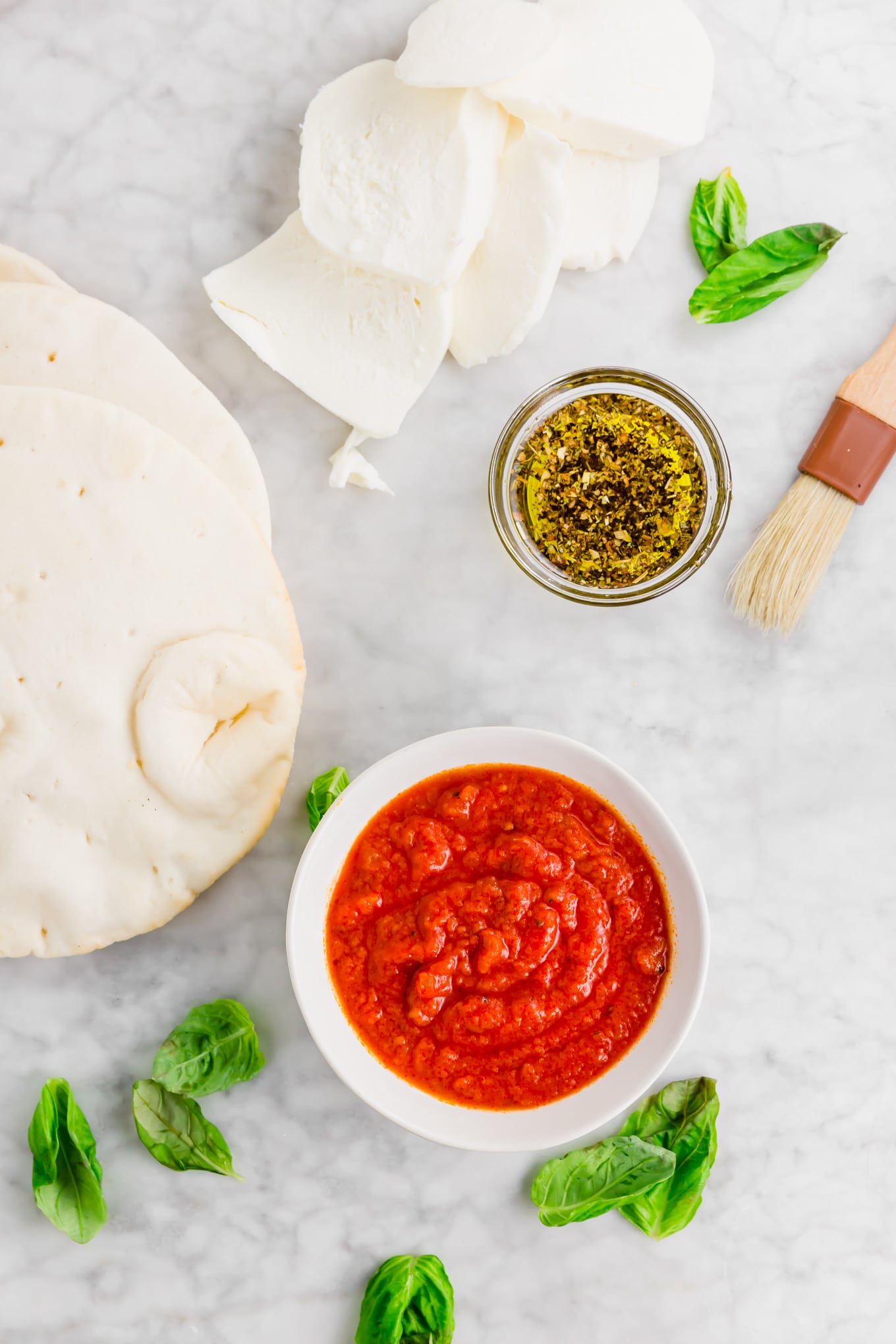 An aerial photo showing the ingredients for making pita pizza: gluten-free pita brea, marinara sauce, fresh mozzarella, olive oil, dried herbs, red pepper flakes, and fresh basil.