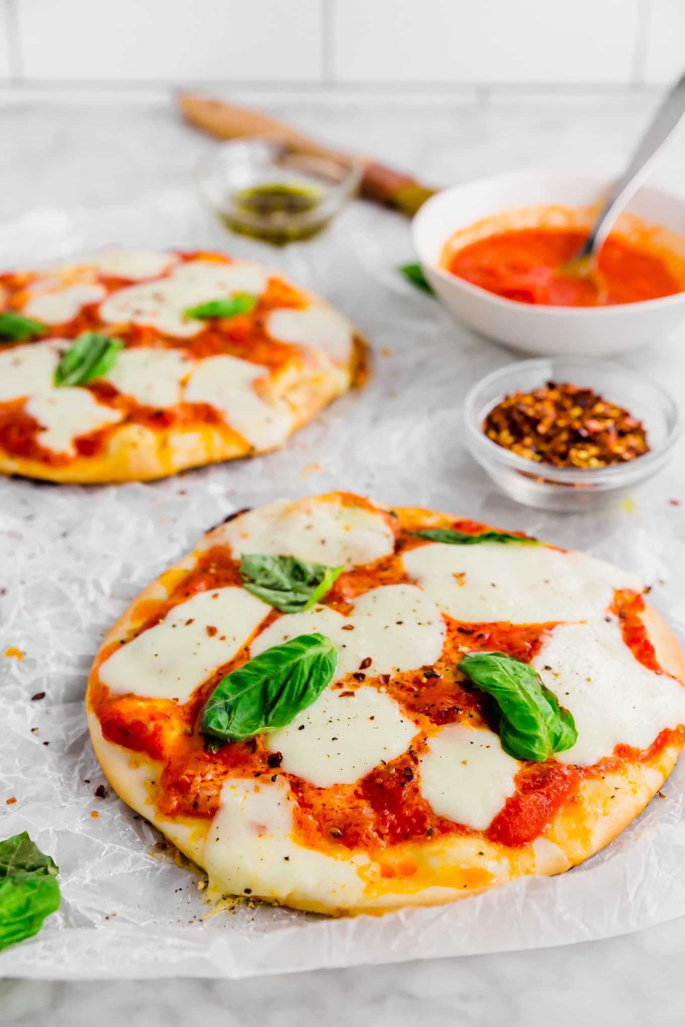 Two pita pizzas with a bowl of marinara sauce and red pepper blacks in the background.