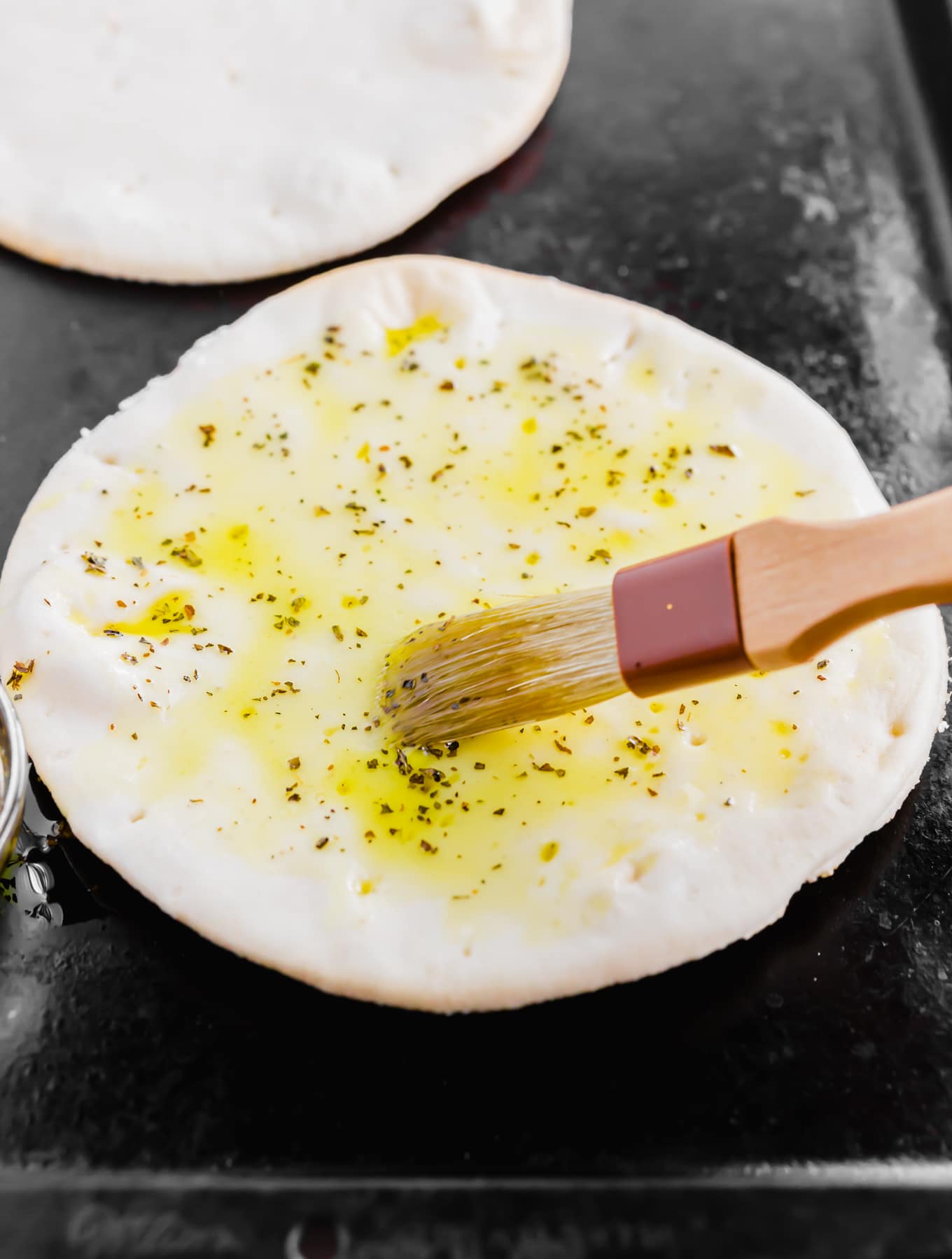 An aerial photo showing a pastry brush spreading an olive oil and herb mixture on gluten-free pita bread.