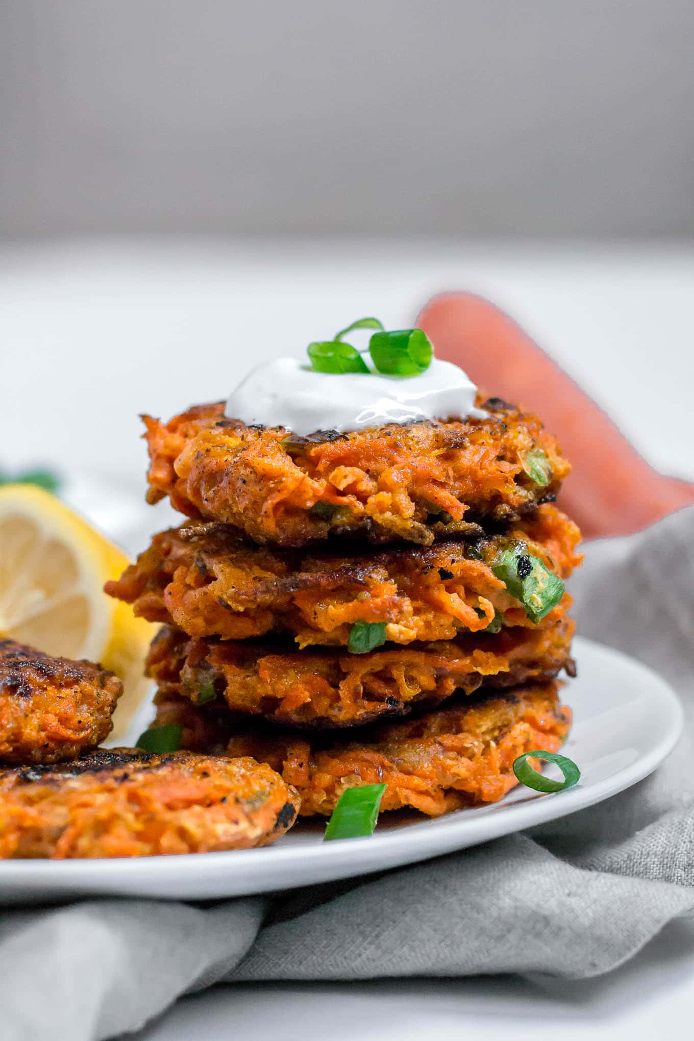A straight on shot of a stack of gluten-free carrot fritters topped with greek yogurt and green onion with a lemon wedge off to the side on a white plate with gray napkin.