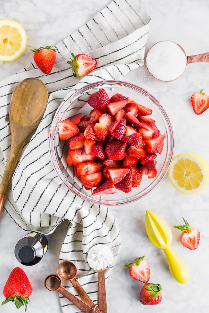 A photo of ingredients for a gluten free strawberry cobbler in bowls - strawberries, lemon, sugar, flour, baking soda, baking powder and vanilla.