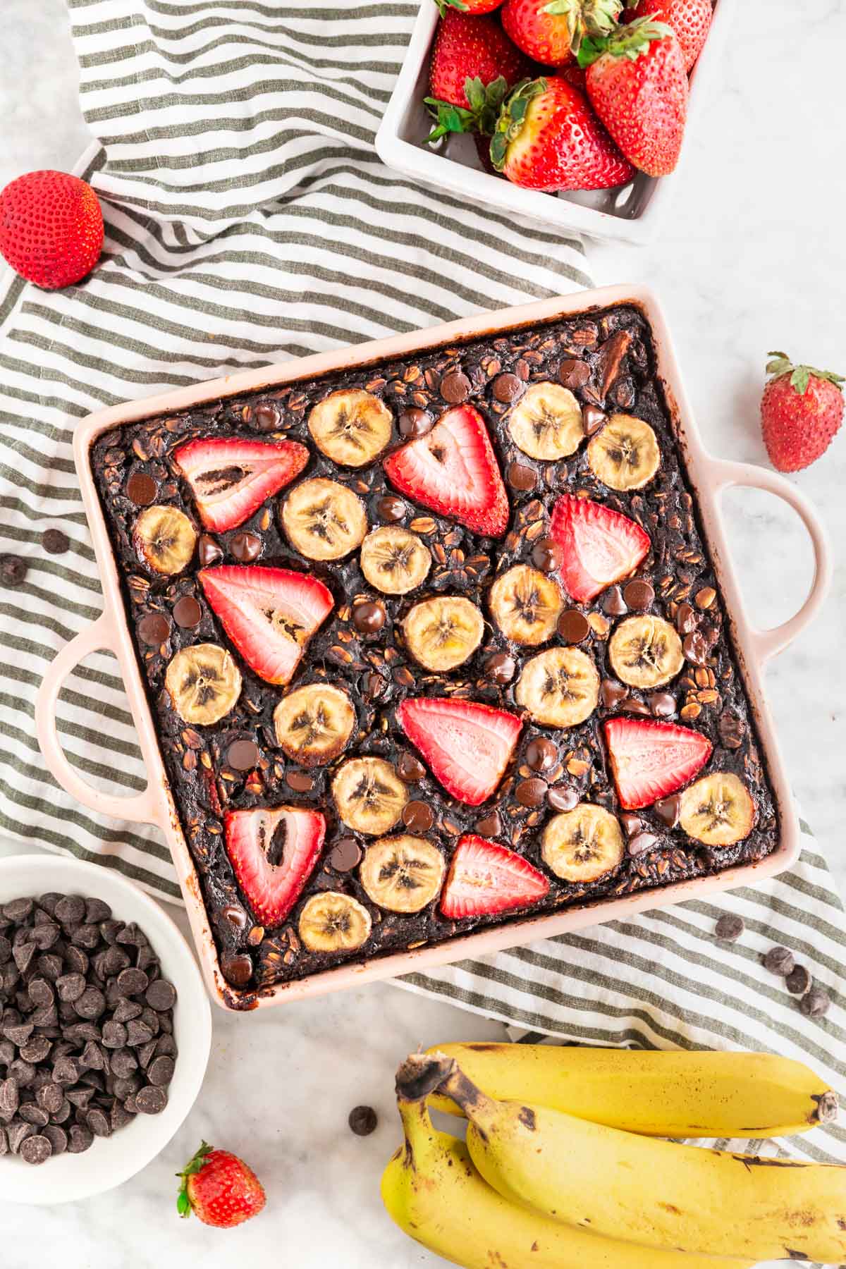 A photo of a baking dish with chocolate strawberry banana baked oatmeal in it with sliced bananas and strawberries on top and a bowl of chocolate chips next to it.
