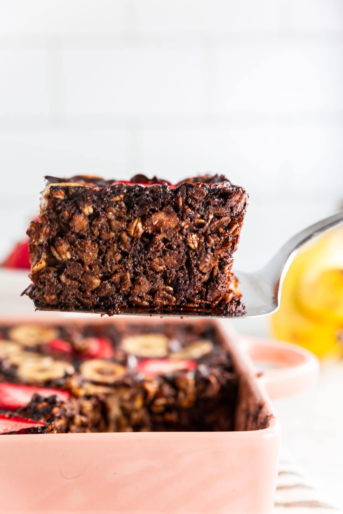 A photo of a slice of chocolate strawberry banana baked oatmeal on a spatula being raised from a baking dish.