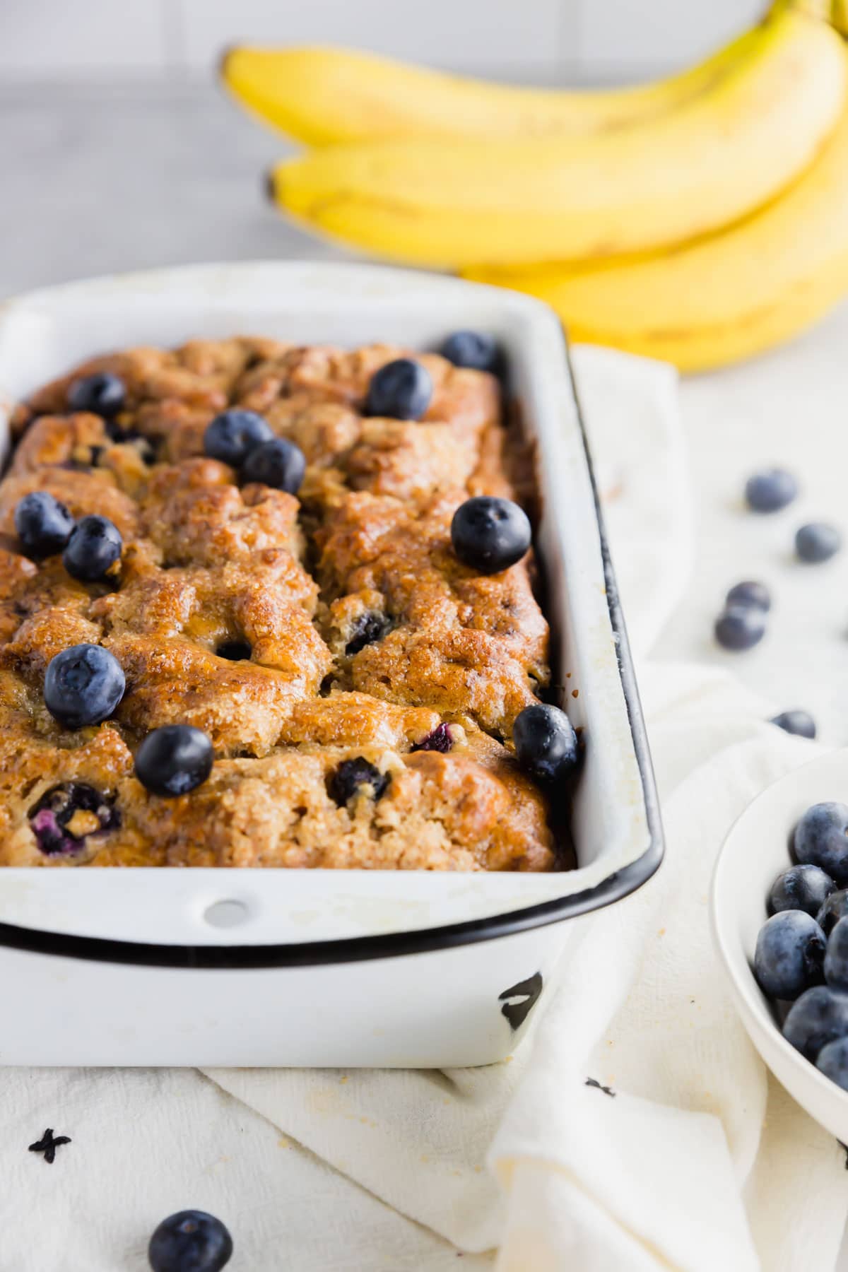 A photo of a gluten-free vegan blueberry banana bread in a white loaf pan with bananas in the background.