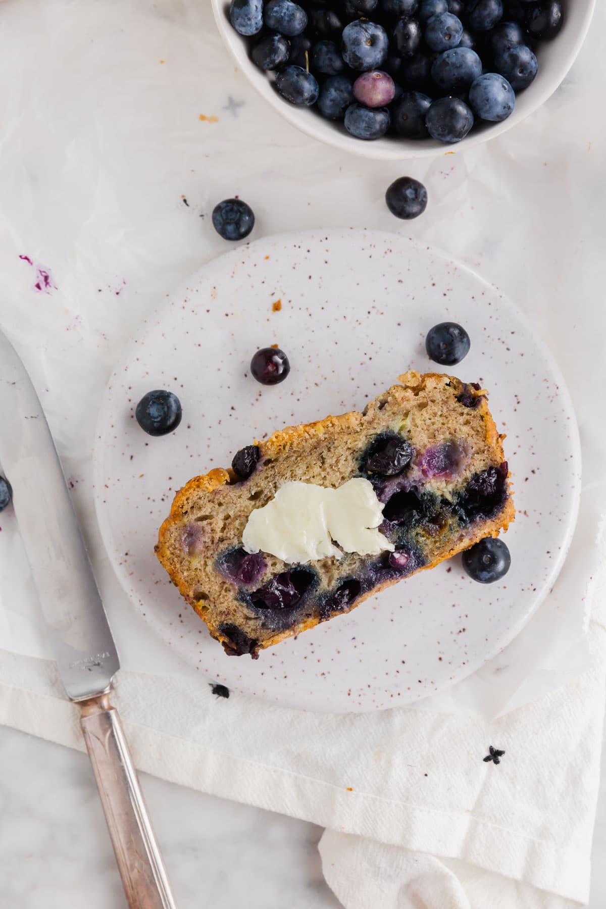 An overhead photo of a slice of gluten-free vegan blueberry banana bread on a small plate topped with vegan butter with a knife next to it and a bowl of blueberries.