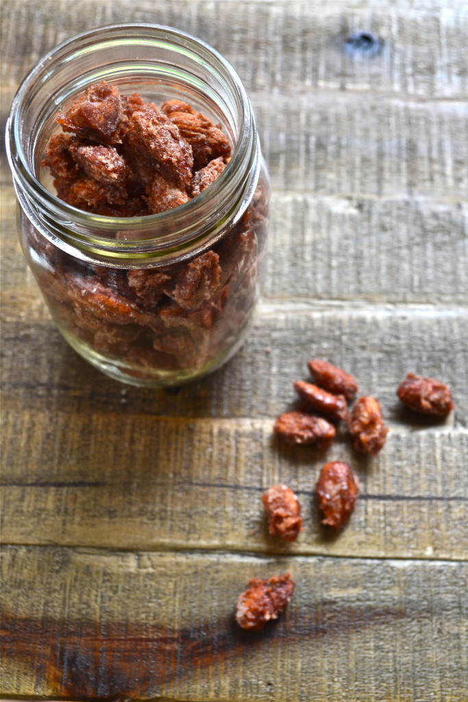 A glass jar filled with candied pecans and candied almonds sits on a rustic wooden surface, with several sugared pecans scattered beside the jar.