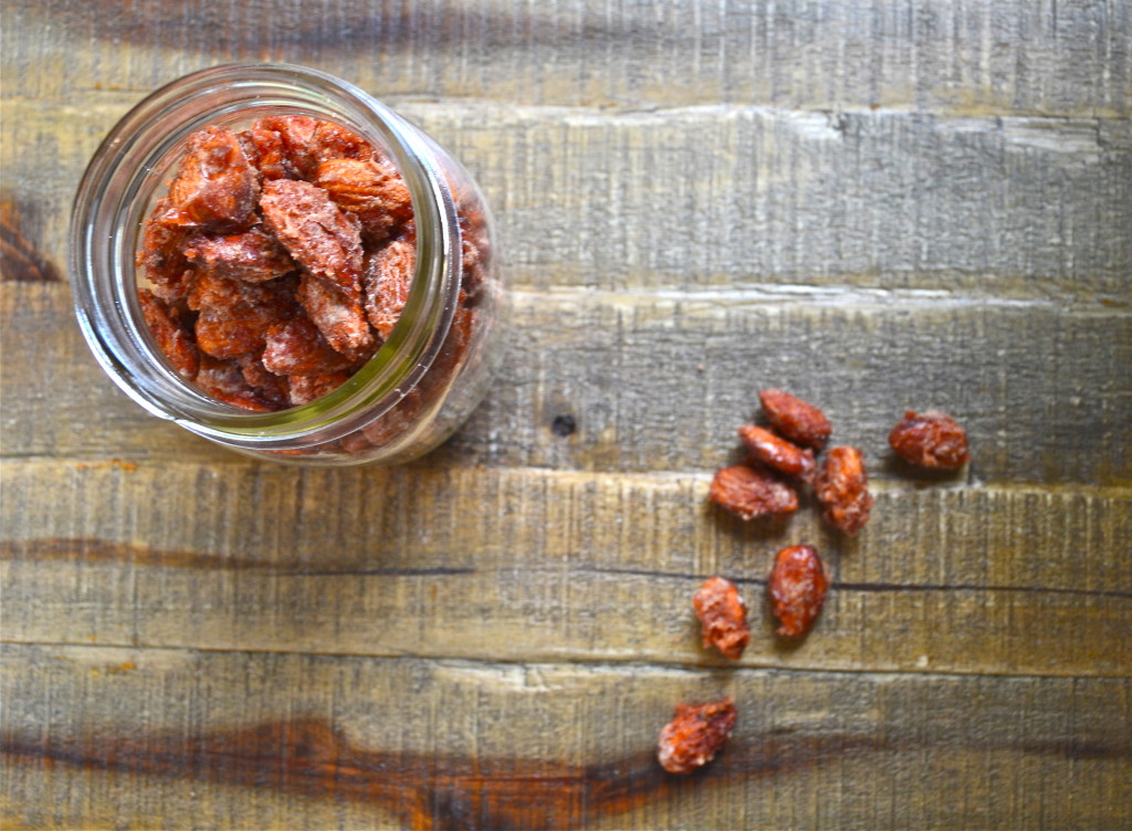 A glass jar filled with sweet candied almonds sits on a wooden surface, with a few candied almonds scattered beside the jar.