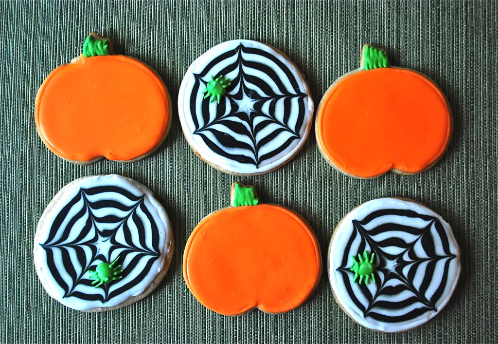 Six decorated cookies, three shaped like bright orange pumpkins and three round ones with black and white spiderweb designs and small green spiders, arranged in two rows on a textured green surface.