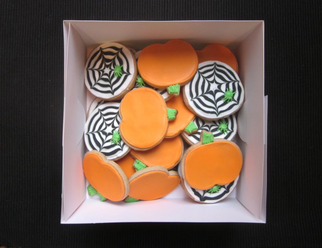 A box filled with decorated cookies, some shaped and iced like orange pumpkins and others round with black and white spiderweb designs and green icing leaves, placed on a dark surface.