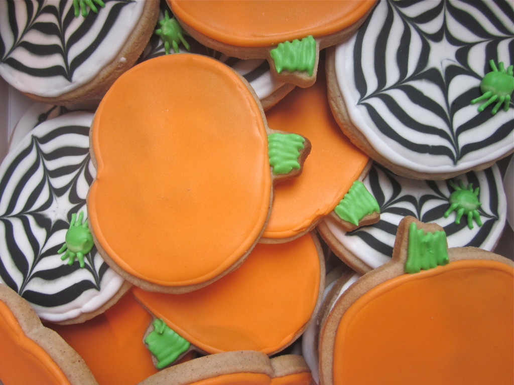 A pile of pumpkin-shaped sugar cookies with orange icing, some decorated with black-and-white spiderweb patterns and small green spiders, and others with plain orange icing and green stems.