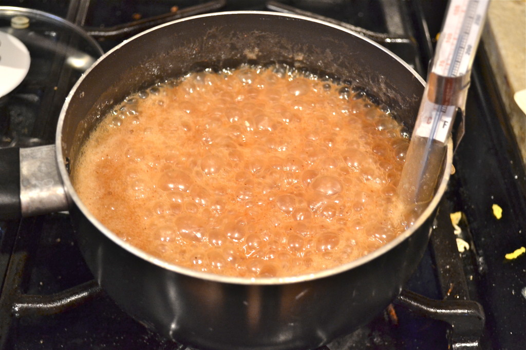 A pot of bubbling brown sugar caramel mixture is boiling on a stovetop, with a thermometer clipped to the side of the pot to measure the temperature.