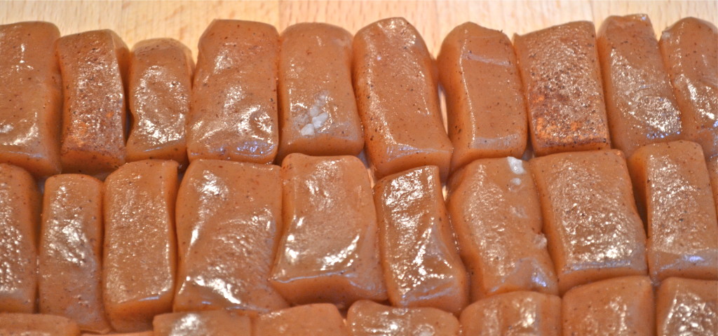 Several rows of rectangular, shiny brown caramel candies are arranged closely together on a wooden surface.
