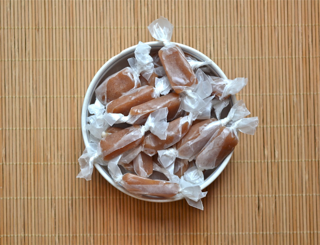 A white bowl filled with individually wrapped caramel candies sits on a bamboo mat background.