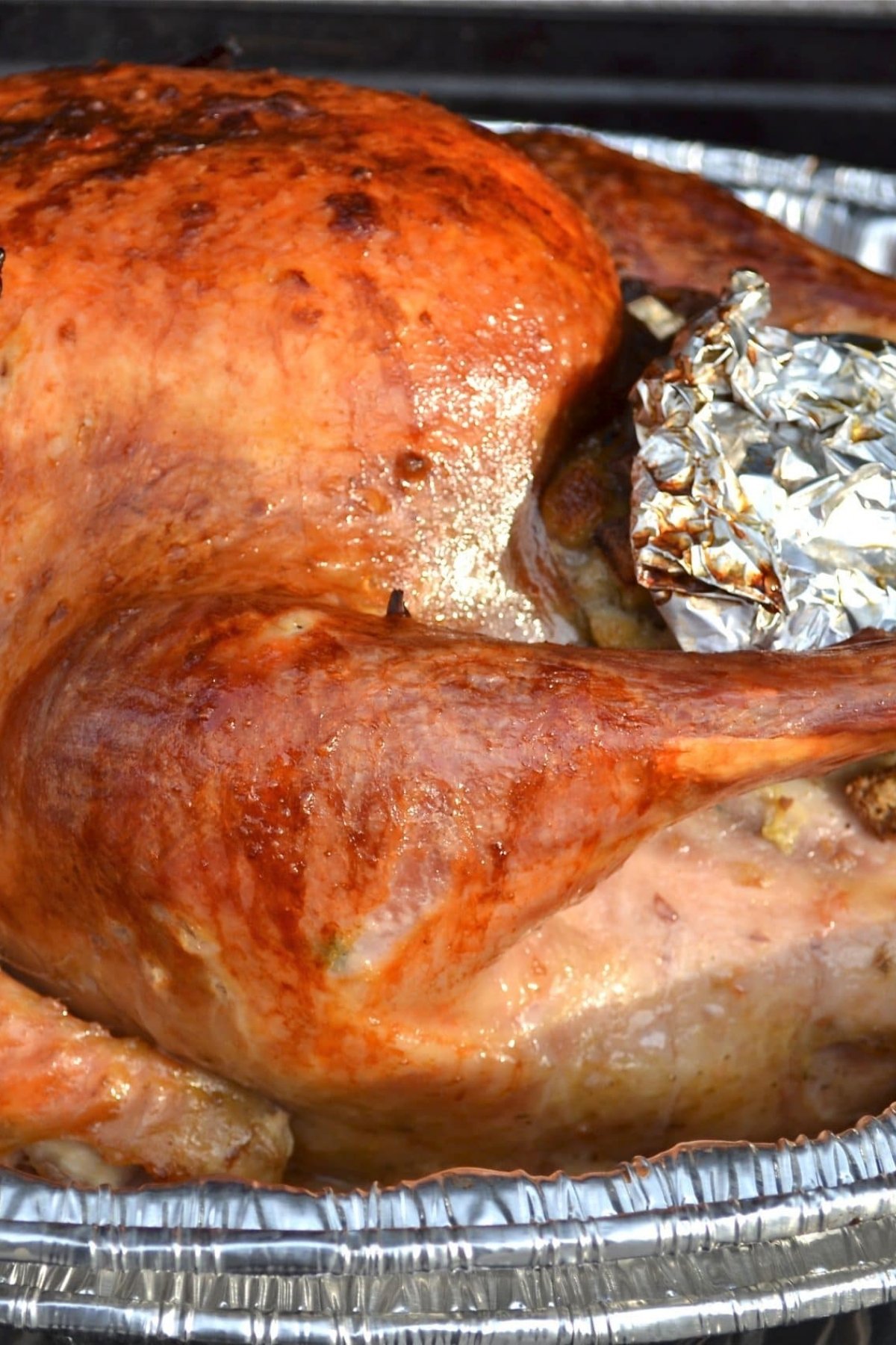 A close-up of a golden-brown grilled turkey in a foil tray, with a piece of aluminum foil covering part of the stuffing near the wing.