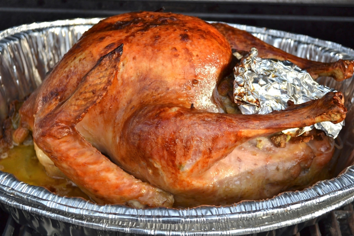 A whole grilled turkey with golden-brown skin sits in a disposable aluminum roasting pan. The cavity is stuffed and partially covered with crumpled foil, while flavorful juices collect at the bottom.