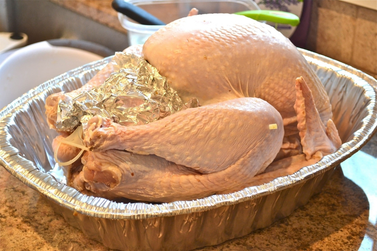 A raw, whole turkey sits in a large aluminum roasting pan on a kitchen counter, ready to become a delicious grilled turkey. The turkey’s cavity is filled with aluminum foil, and kitchen tools are visible in the background.