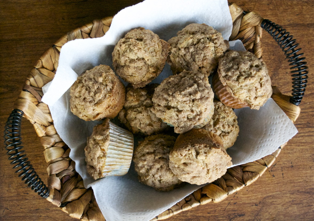 A woven basket lined with a white paper towel holds a pile of homemade Cinnamon Muffins with Streusel Topping {Gluten-Free}, each muffin boasting a crumbly, golden-brown top, sitting on a wooden surface.