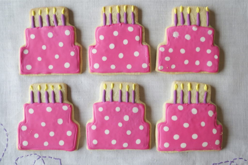 Six Gluten-Free Birthday Cake Sugar Cookies are decorated as pink cakes with white polka dots, yellow candles, and purple candle bases, arranged in two rows on a white surface.