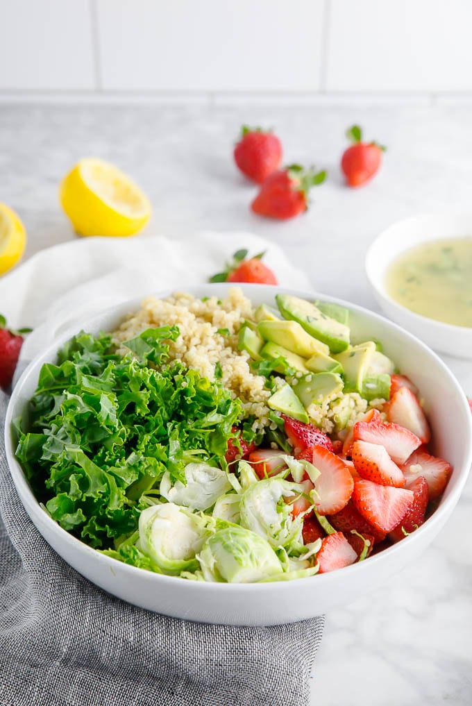 A bowl of kale, brussels sprouts, quinoa, avocado, and strawberries in a bowl ready to mix with lemon vinaigrette.