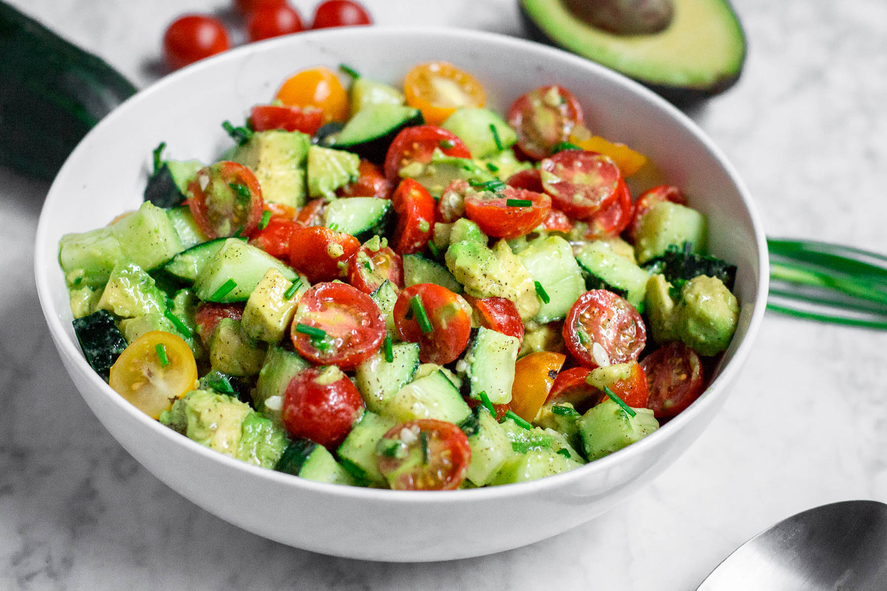 A white bowl filled with diced cucumbers, tomatoes, and avocado in a lemon dressing with chopped chives on top.