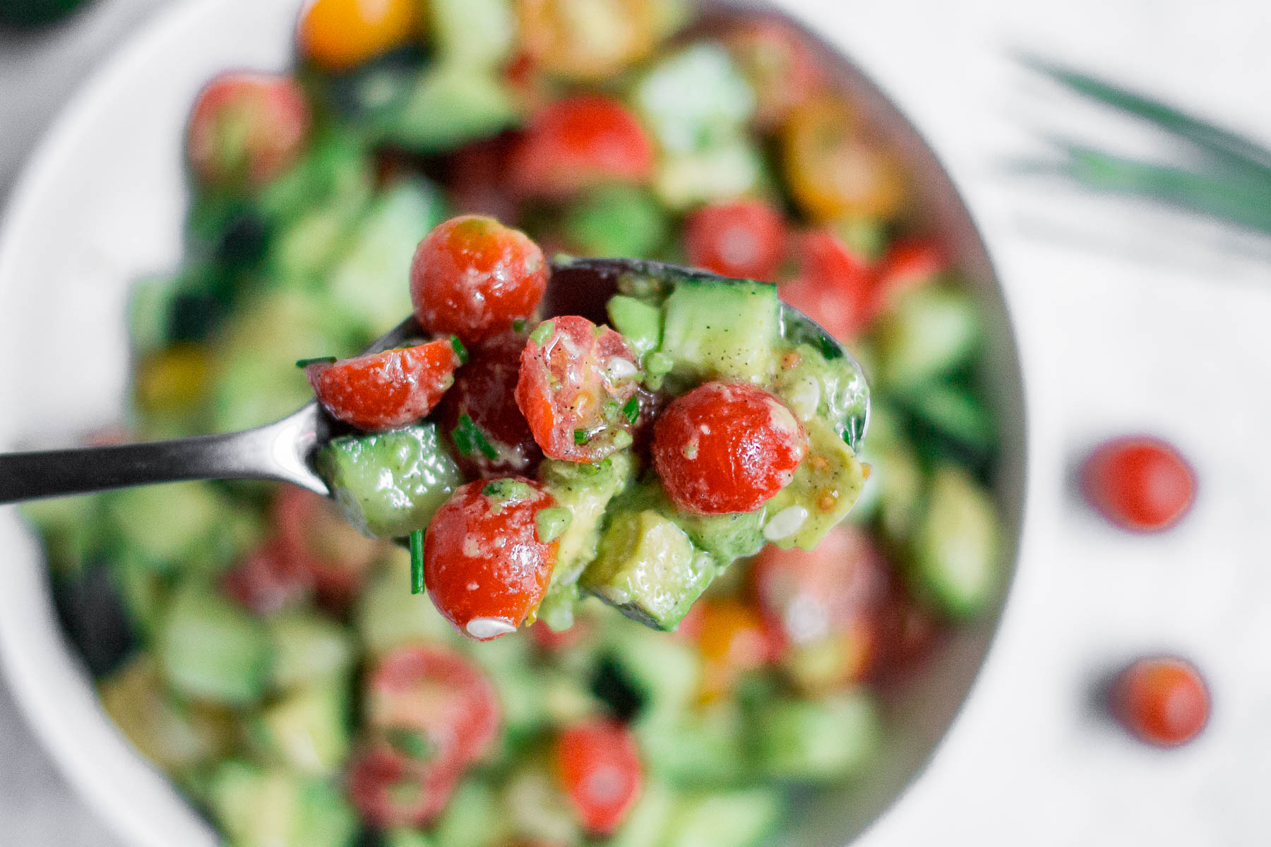 A close-up of a spoon holding a fresh Cucumber Tomato Avocado Salad with cherry tomatoes, cucumber, and avocado, with a blurred bowl of the same salad in the background.