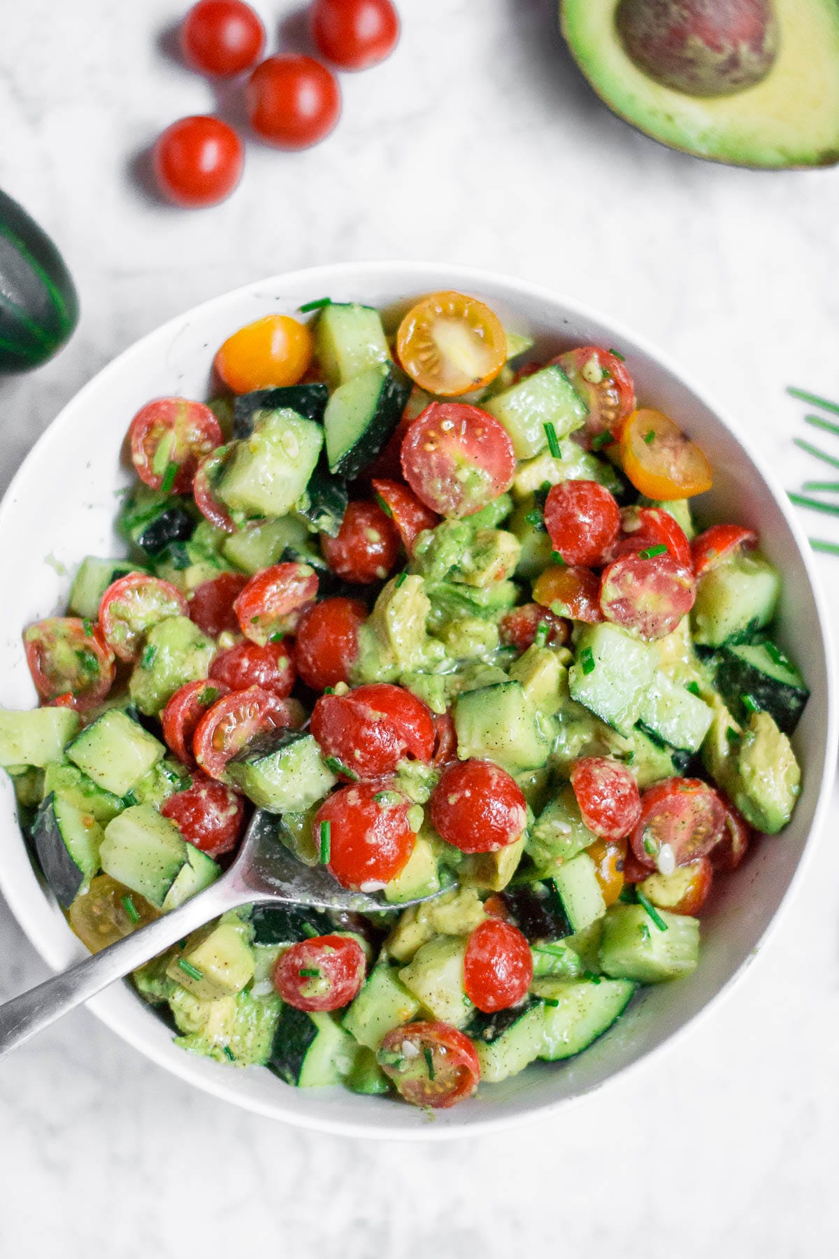 A bowl of Cucumber Tomato Avocado Salad with sliced cherry tomatoes, diced avocado, cucumber, and herbs, mixed together. A spoon is in the bowl, with whole cherry tomatoes and half an avocado on a white surface.
