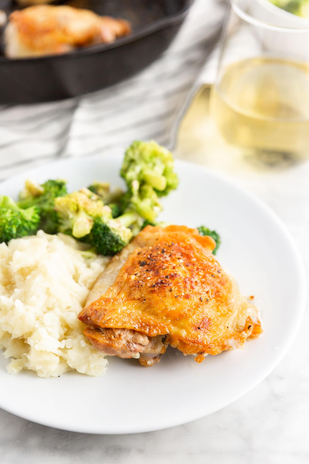 A dinner plate with roasted chicken thighs, mashed potatoes and steamed broccoli with a cast iron skillet and a glass of wine in the background.