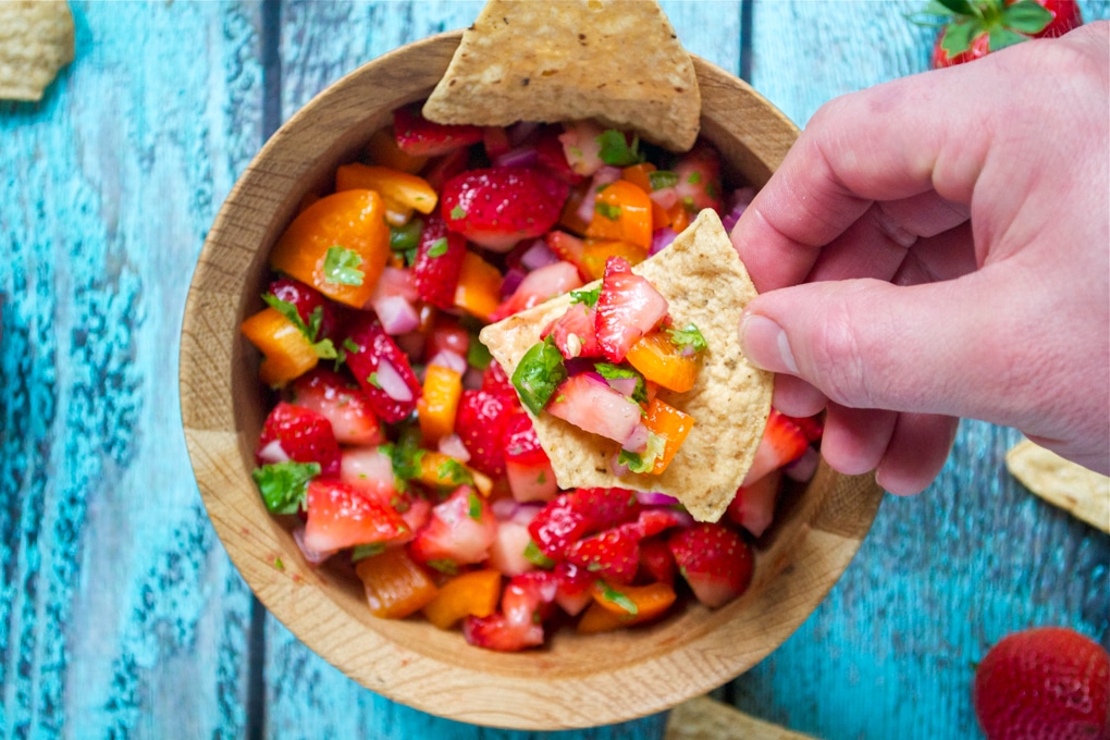 A hand holding a tortilla chip topped with fresh strawberry salsa over a wooden bowl filled with chopped strawberries, orange bell pepper, red onion, and cilantro.