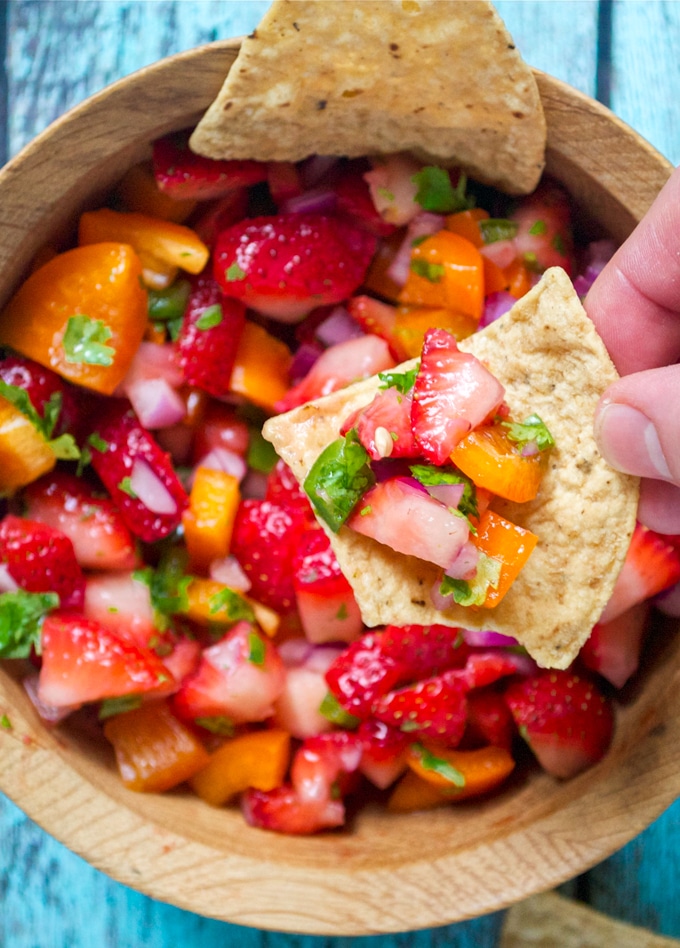 Close-up of a tortilla chip being dipped into strawberry salsa with chopped strawberries, orange bell pepper, red onion, jalapeño, and fresh cilantro in a wooden bowl.