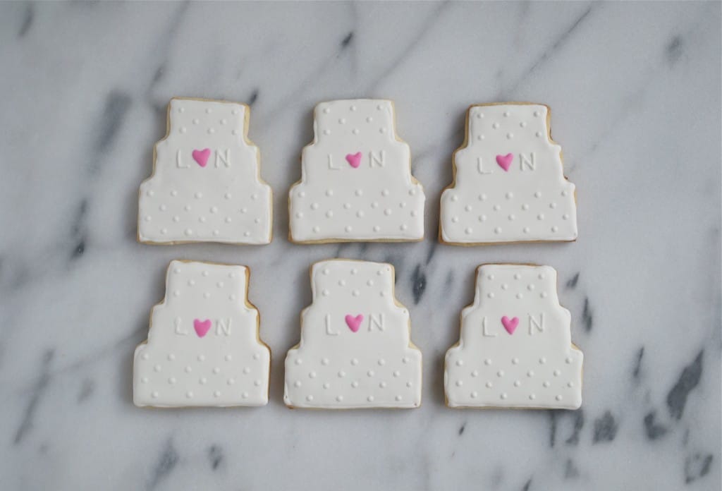 Six wedding cake sugar cookies decorated with royal icing on a marble table.
