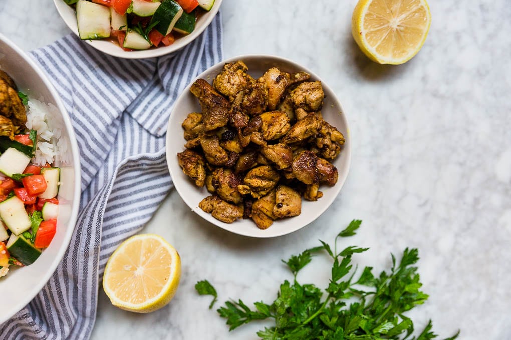 A bowl of spiced chicken thighs for easy mediterranean chicken bowls with lemon wedges, tomato cucumber salad and fresh parsley.