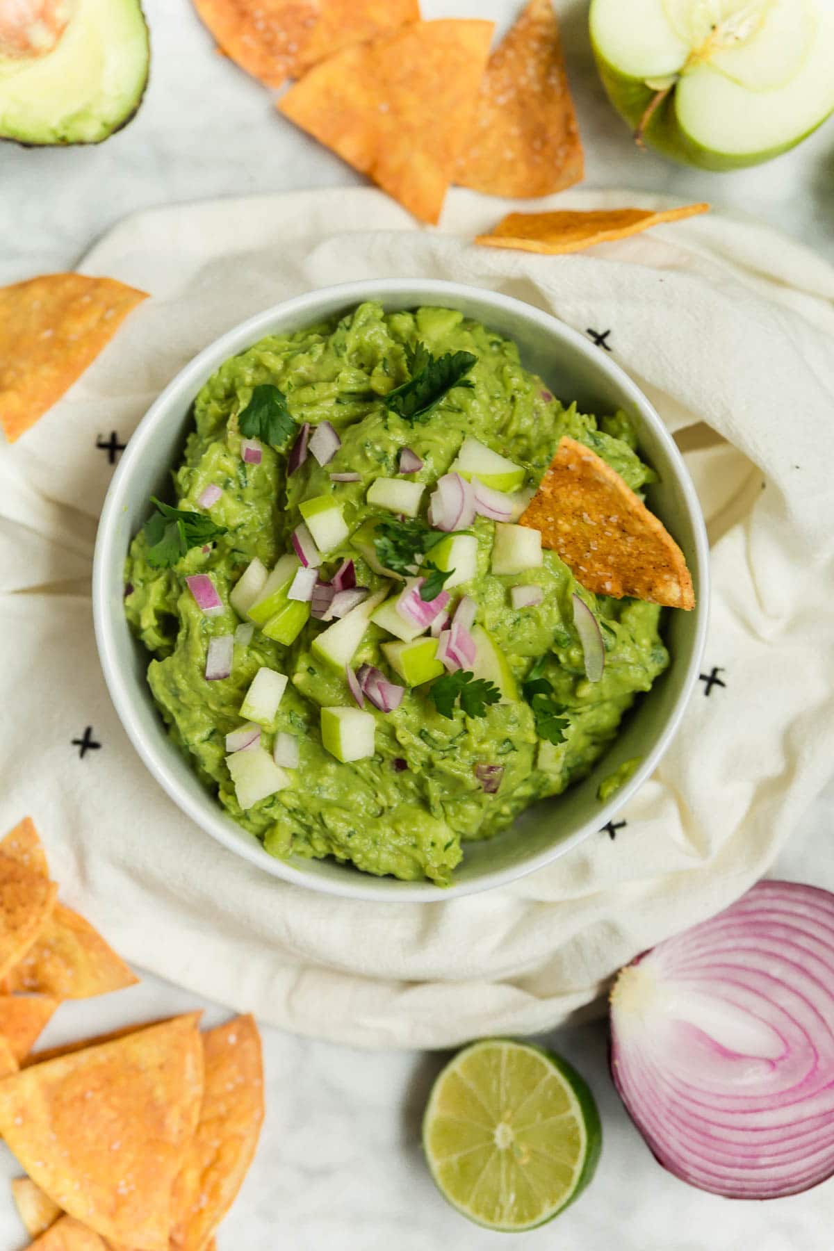 A bowl of green apple guacamole with baked tortilla chips, red onion, and limes.