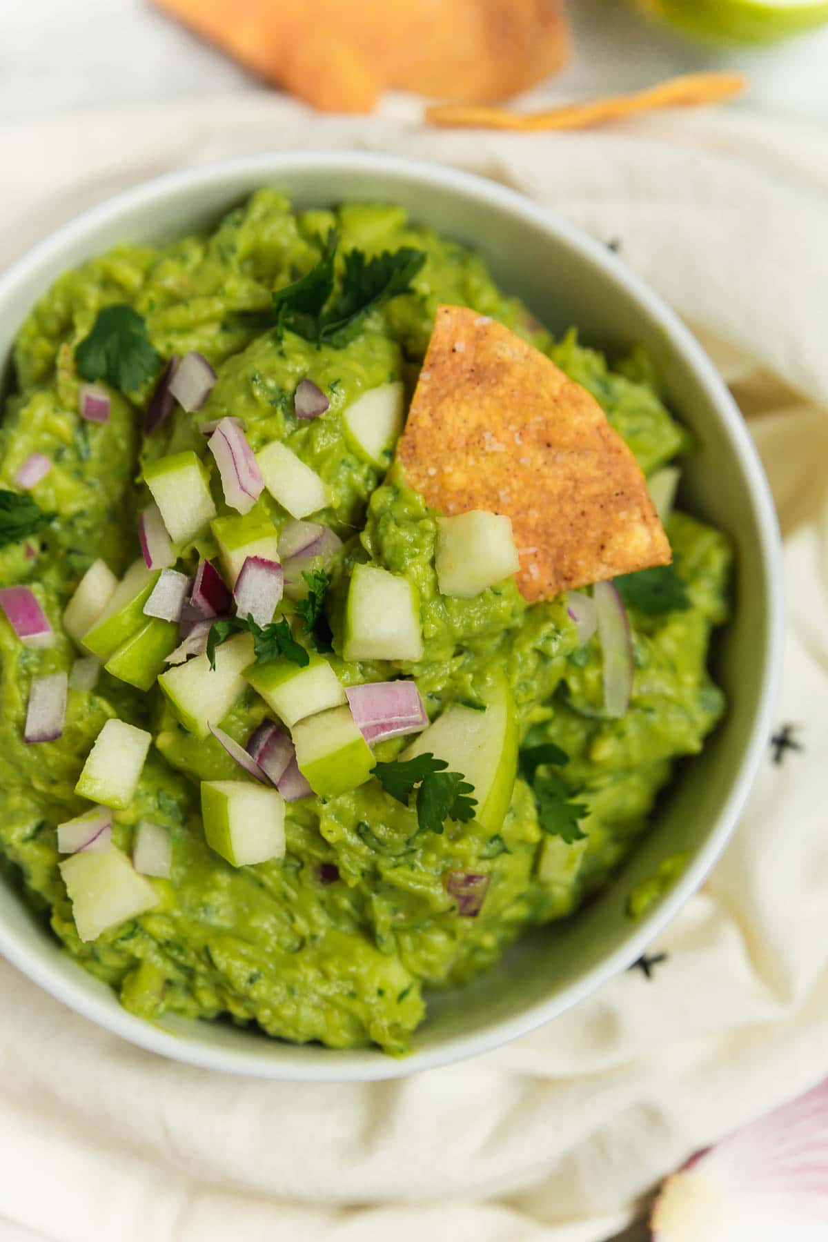 A bowl of guacamole with apple, red onion, cilantro and lime with a tortilla chip.
