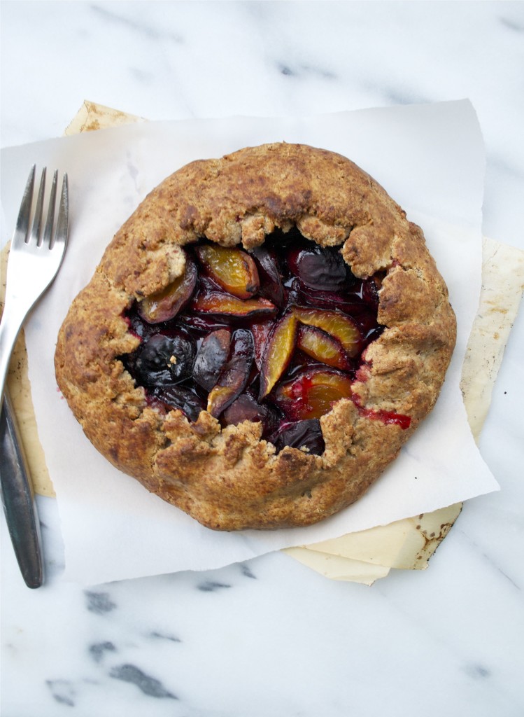A Rustic Italian Plum Galette with a golden brown crust and visible plum filling sits on parchment paper. A fork and knife rest beside it on a white marble surface.