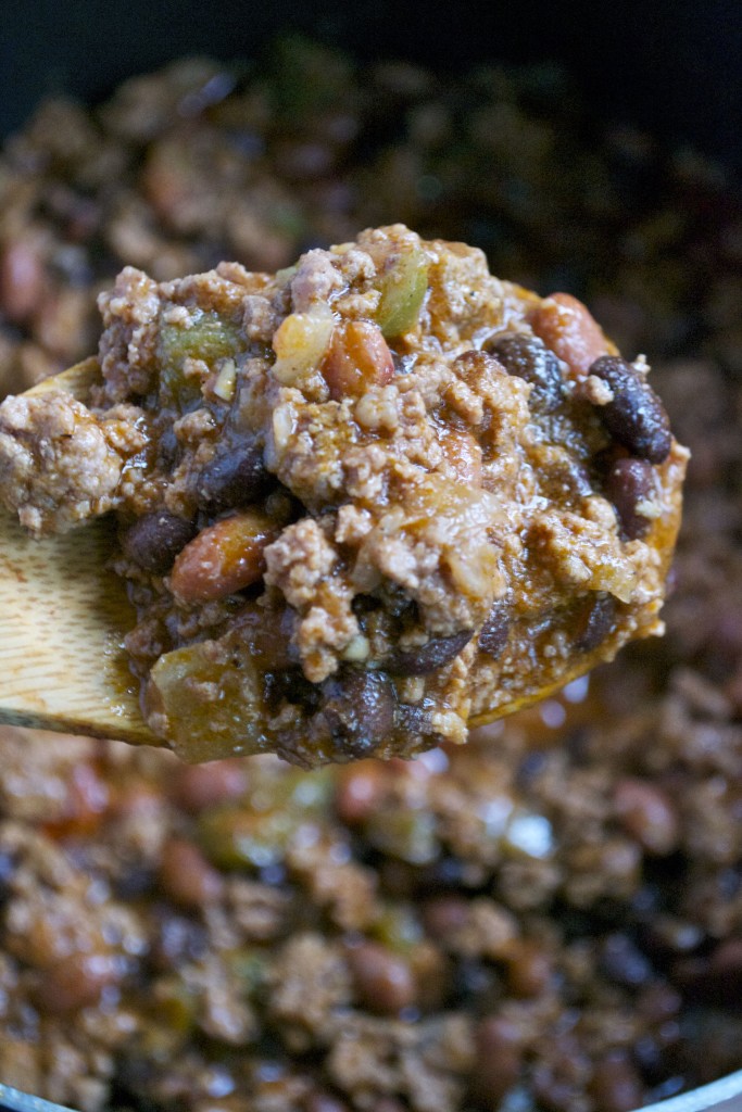 A close-up of a wooden spoon lifting a hearty Gluten-Free Beef Chili made with ground meat, beans, tomatoes, and peppers, with more chili visible in the background.