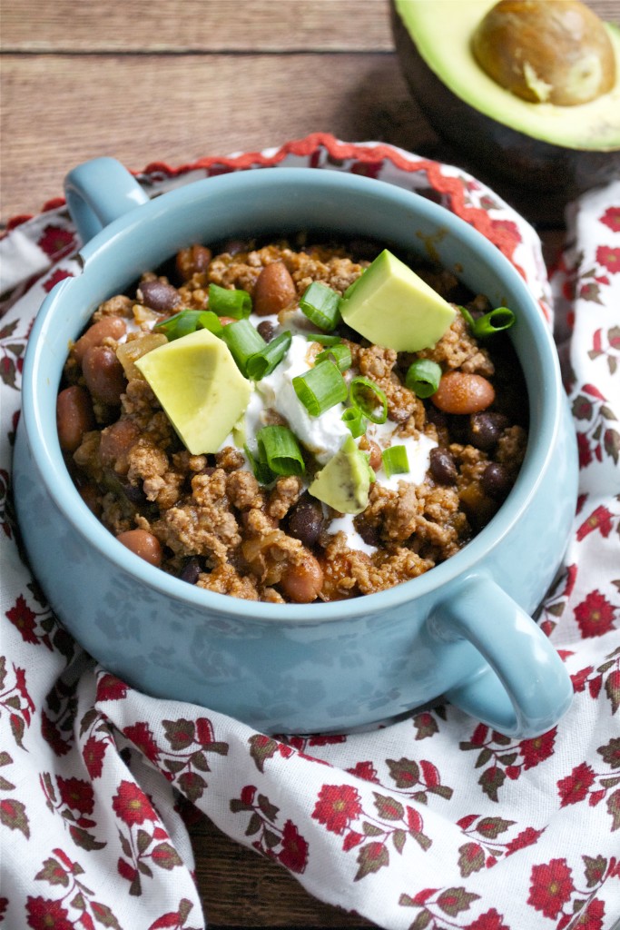 A blue bowl filled with Quick and Easy Beef Chili, topped with sour cream, diced avocado, and chopped green onions sits on a floral-patterned cloth with a halved avocado in the background.