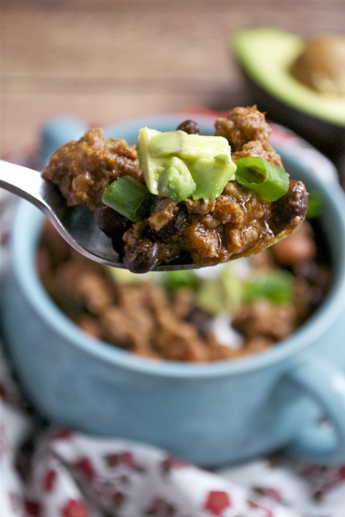 A spoonful of Gluten-Free Beef Chili with ground meat, black beans, green onions, and diced avocado is held above a blue bowl; a halved avocado sits in the blurred background.