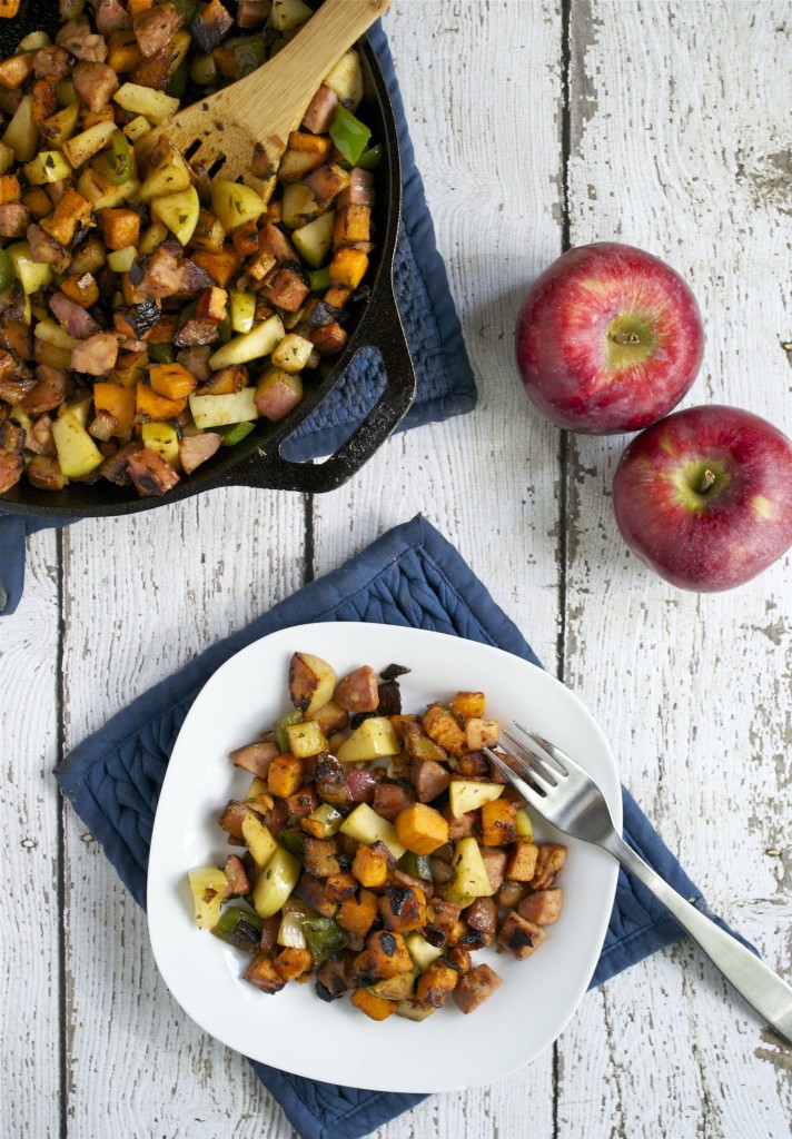A white plate with colorful Apple and Sausage Breakfast Hash—diced apples, sweet potatoes, sausage, and herbs—sits on a blue mat next to a fork. A cast iron skillet with more hash and two red apples rest on a white wooden table.