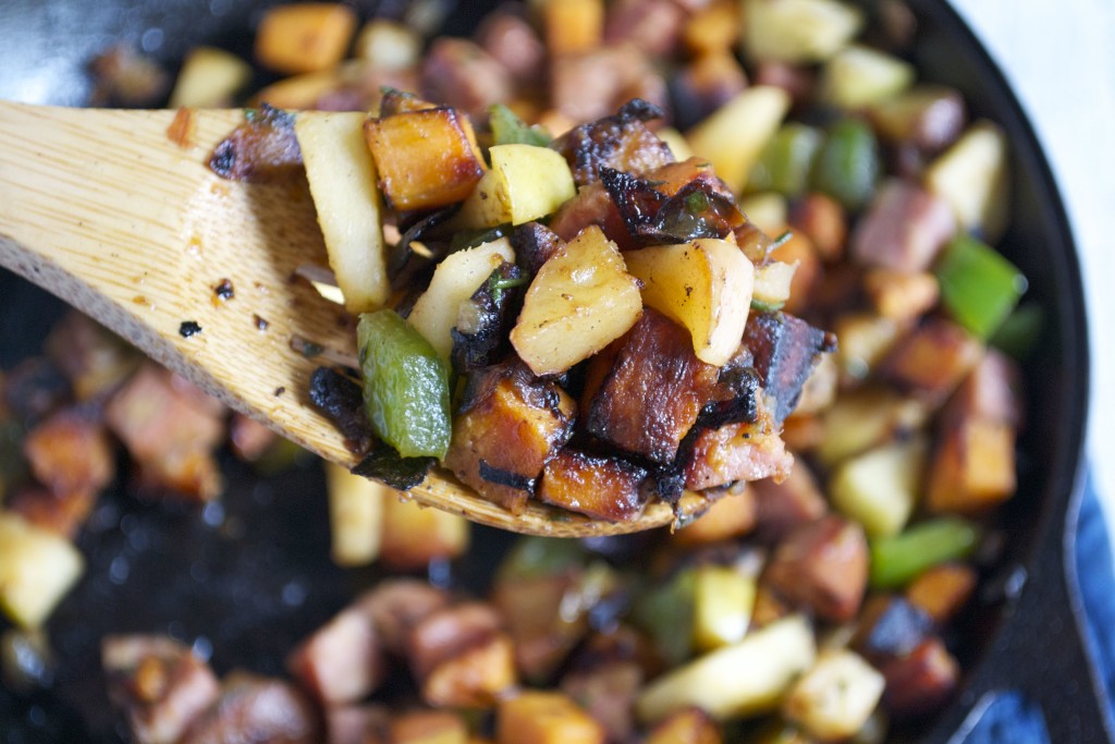A close-up of a wooden spoon lifting a mix of sautéed potatoes, sweet potato, green bell peppers, onions, and sausage from a skillet. The vegetables and sausage are browned and cooked for an Apple and Sausage Breakfast Hash.