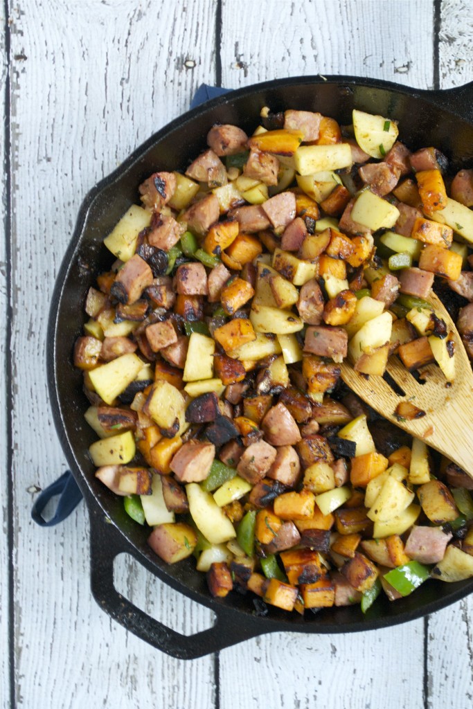A cast iron skillet filled with Sweet Potato, sausage, green peppers, and onions sits on a white wooden surface. A wooden spatula rests in the skillet, mixing the browned and colorful ingredients for a delicious Apple and Sausage Breakfast Hash.