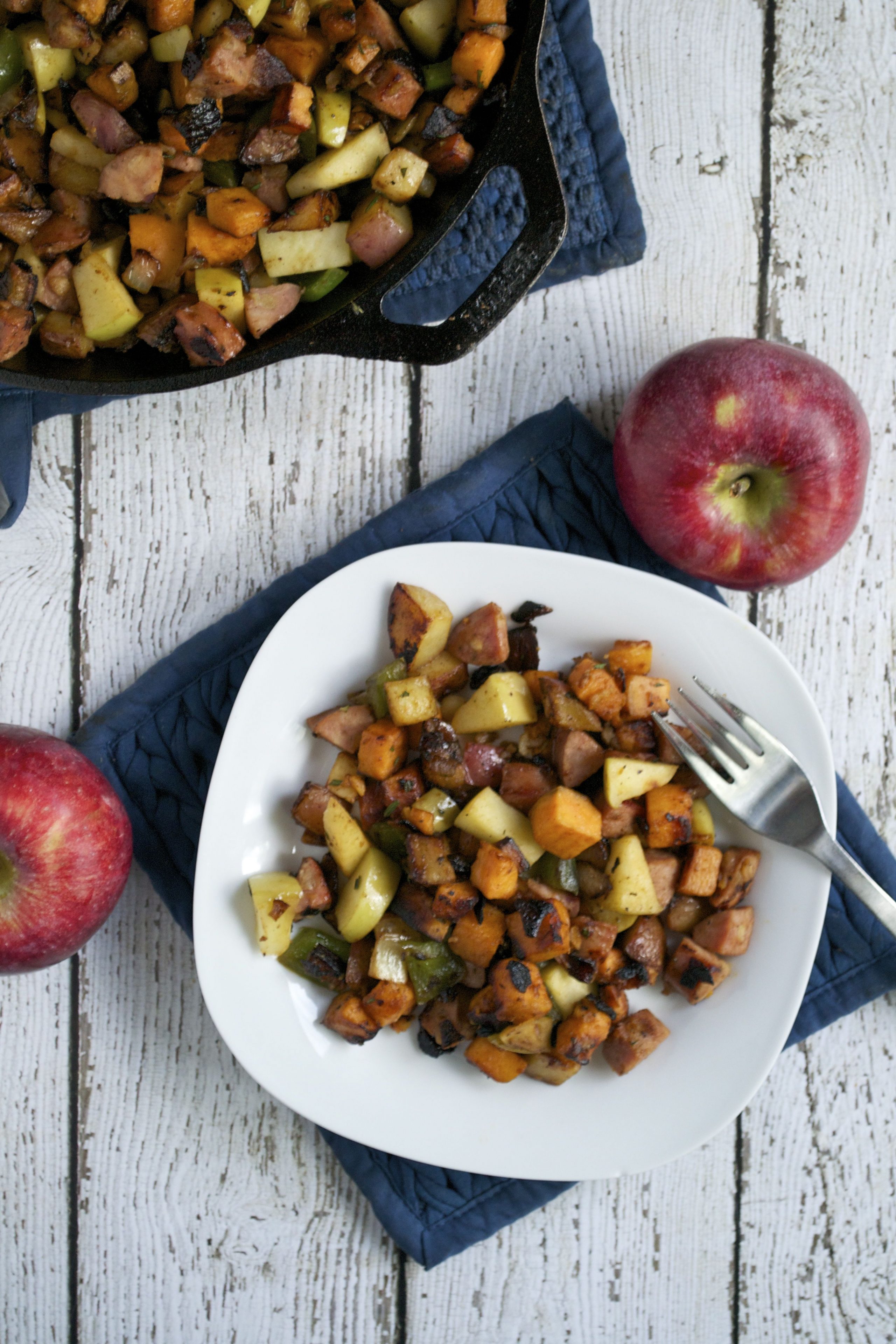 A white plate with Sweet Potato, Apple and Sausage Breakfast Hash sits on a blue quilted mat beside a fork. Two whole red apples and a skillet with more hash rest on a rustic white wooden table.