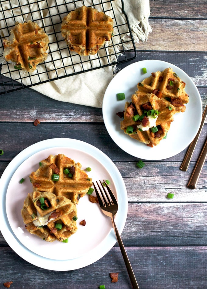 An overhead view of mashed potato waffles on two plates topped with green onion and bacon with additional waffles on a cooling rack.
