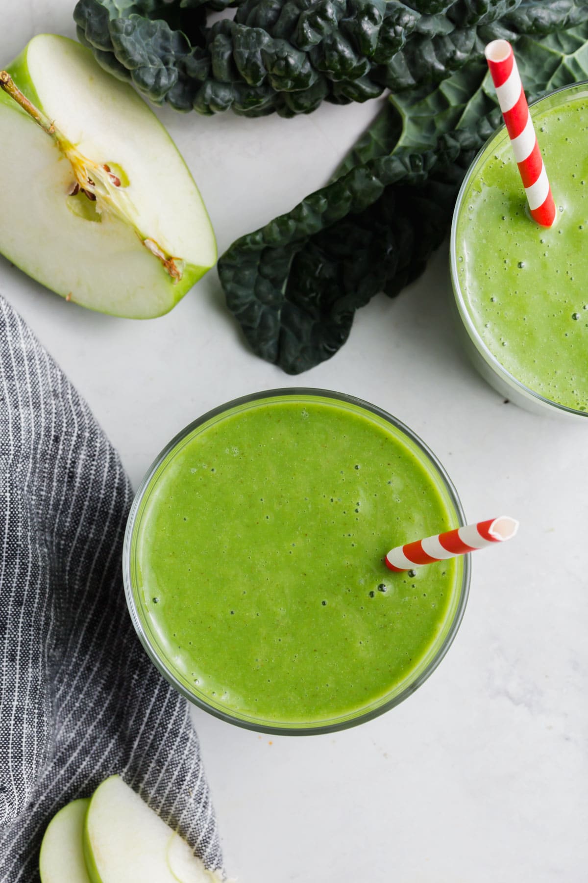 An overhead photo of two glasses of kale apple green smoothies with kale and apples on the table.