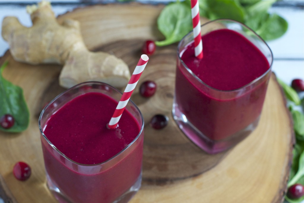 Two glasses of bright ruby red cranberry beet smoothie with red-striped straws, set on a wooden board with fresh ginger, spinach leaves, and cranberries in the background.